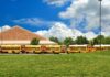 Waymo Robotaxi Flouts LAW – Kids Endangered! A row of yellow school buses parked in front of a school building under a cloudy sky
