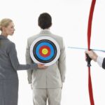 A woman places a target on a mans back while another man aims a bow and arrow at it