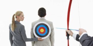 A woman places a target on a mans back while another man aims a bow and arrow at it