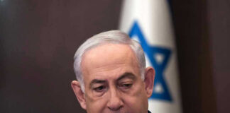 Man in suit with Israeli flag in background.
