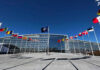 Trump and Kazakhstan Unveil $17 Billion Partnership Flags outside NATO headquarters building under clear blue sky.
