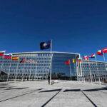 Flags outside NATO headquarters building under clear blue sky.
