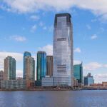 A modern city skyline featuring tall buildings along a waterfront under a cloudy sky