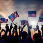 Silhouetted crowd holding American flags against a sunset sky