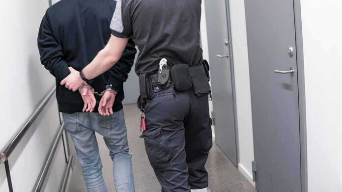 shutterstock_1082126789-1 Security officer escorts handcuffed person down hallway