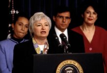 Federico Peña stands behind a woman speaking at a podium with the presidential seal