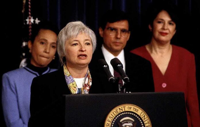 Federico Peña stands behind a woman speaking at a podium with the presidential seal